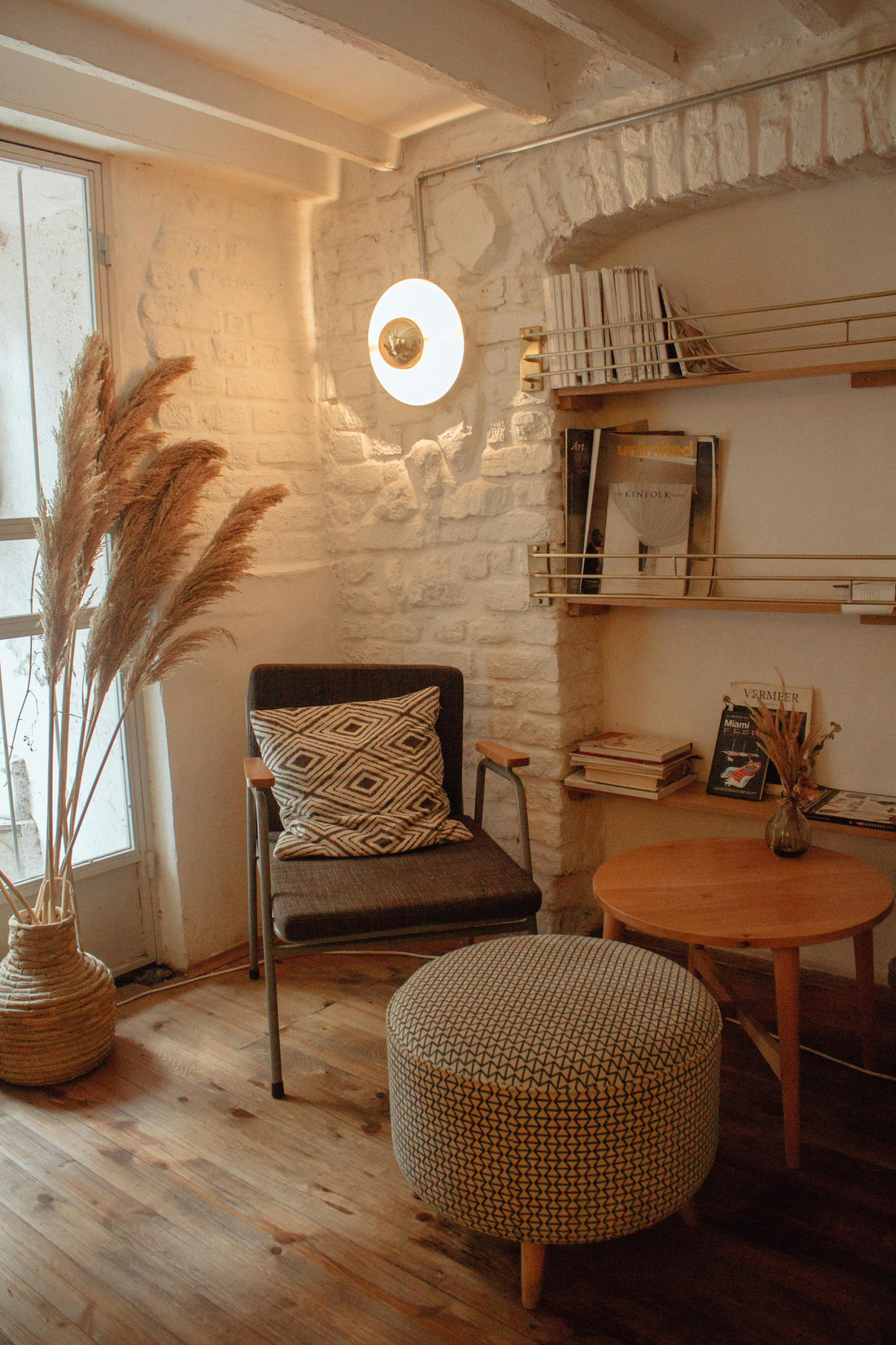 Cozy reading nook with a wooden chair, patterned cushion, round ottoman, and a warm wall light, set against a rustic brick wall with shelves.