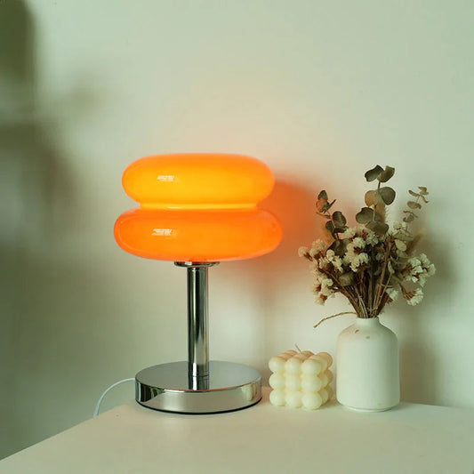 Modern table lamp with a soft orange glow, placed next to dried flowers and a decorative candle on a minimalist white surface.