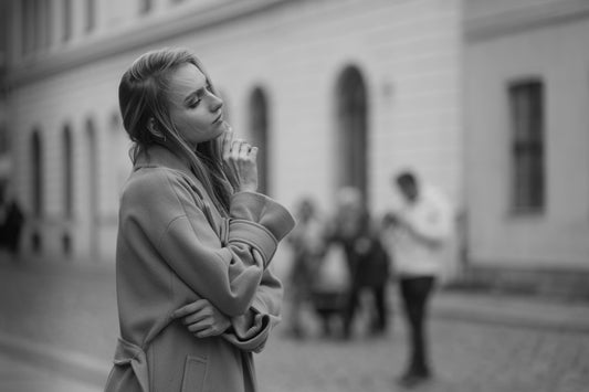 A black-and-white image of a woman standing outdoors, deep in thought, with blurred figures in the background.