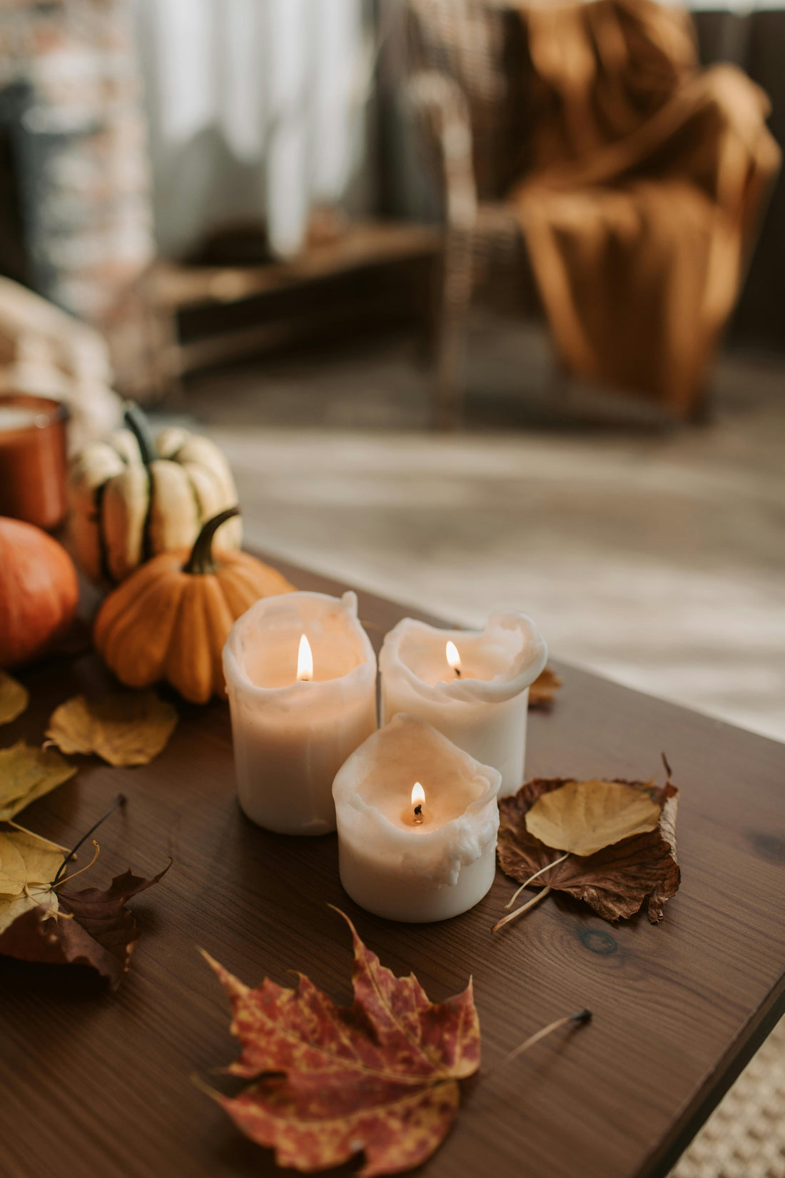 Lit candles on a wooden table surrounded by autumn leaves and small pumpkins, creating a cozy fall atmosphere.